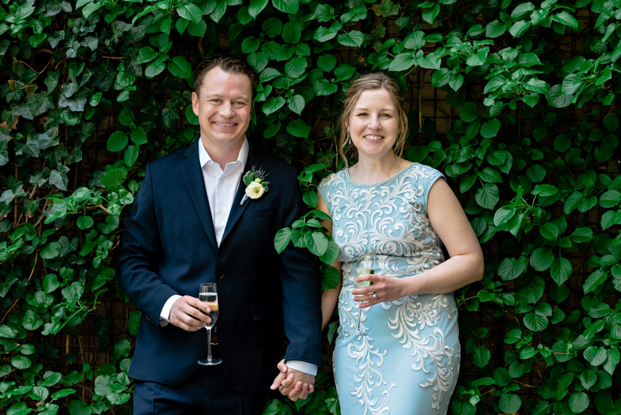 A bride and groom hold hands and stand against an ivy wall.